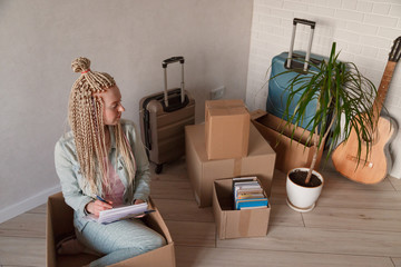 Beautiful woman sitting in box in casual clothes is packing her stuff into the boxes, making notes