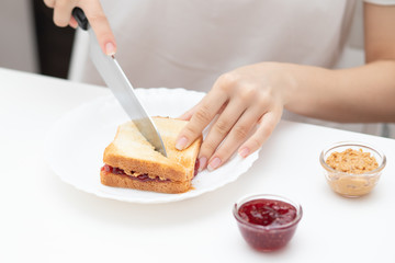 A young woman cuts an American sandwich with jam and peanut butter, prepares to eat it for breakfast in the morning at home.