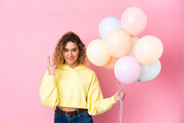 Young blonde woman with curly hair catching many balloons isolated on pink background pointing with the index finger a great idea