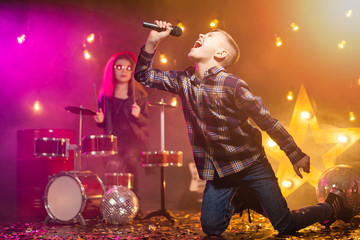 Kids pretending to be a rock band play and sing at studio or stage. Boy singing and girl paying on drums