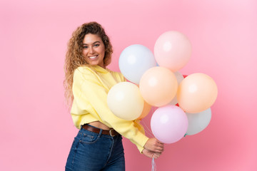 Young blonde woman with curly hair catching many balloons isolated on pink background