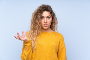 Young blonde woman with curly hair isolated on blue background making doubts gesture