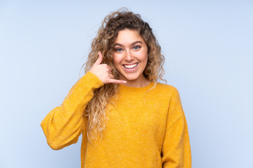 Young blonde woman with curly hair isolated on blue background making phone gesture. Call me back sign