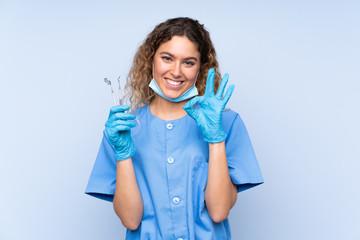 Young blonde woman dentist holding tools isolated on blue background showing an ok sign with fingers