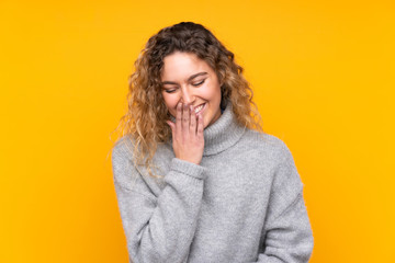 Young blonde woman with curly hair wearing a turtleneck sweater isolated on yellow background smiling a lot