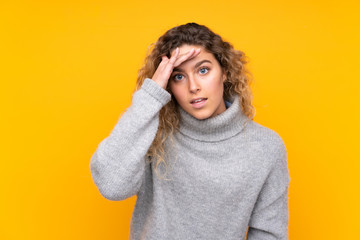 Young blonde woman with curly hair wearing a turtleneck sweater isolated on yellow background looking far away with hand to look something
