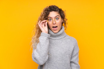 Young blonde woman with curly hair wearing a turtleneck sweater isolated on yellow background with glasses and surprised