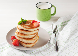 Russian traditional cuisine, wheat flour pancakes on a light plate on a white table, pancakes with fresh strawberries and a mint leaf, a green speckled mug with milk