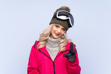 Skier teenager girl with snowboarding glasses over isolated blue background points finger at you