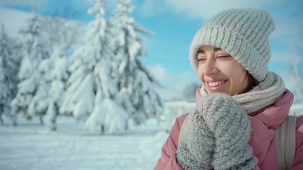 slow motion dolly shot beautiful young woman in warm pink parka and knitted hat and scarf posing in snowy winter park at frozzy sunny day on snowy fir and blue sky background and heating hands by