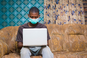 young black man sitting at home on a couch, wearing a face mask, using his laptop