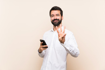 Young man with beard holding a mobile happy and counting three with fingers - obrazy, fototapety, plakaty