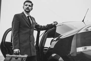 Portrait of a young bearded businessman of thirty. Holding a leather bag in his hand. In a business style, stands at the company car, looking to the side. Against the sky. In creative tinting