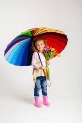 little blonde girl is smiling, standing in rubber boots holding spring flowers and a multi-colored umbrella on a white background