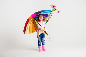 little blonde girl is smiling, standing in rubber boots holding spring flowers and a multi-colored...