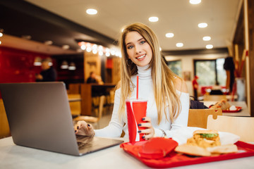 Stylish blonde girl student is learning with a laptop in a cafe,eating fast food and drinking a...