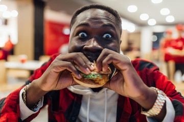 A portrait of young african man eating a burger in street food cafe. Fast food eating time.