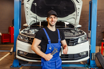 male technician showing thumb up and holding wrenches while smiling at the camera in the service station