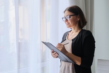 Mature woman teacher taking notes, standing near window in office