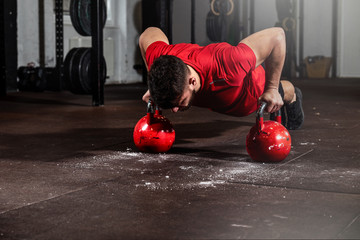 Young strong fit sweaty muscular man with big muscles doing push ups on two big old heavy kettlebells with his hands for hard core cross workout training on the gym floor real people