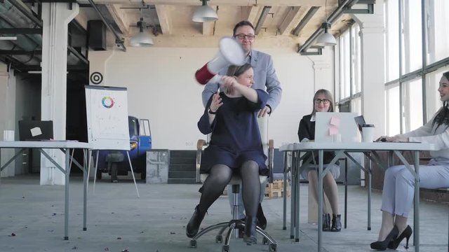Colleagues congratulate the woman manager, roll her in an office chair, blow up crackers with confetti. Woman singing in a megaphone and dancing with a business team. Corporate. Office. Celebration