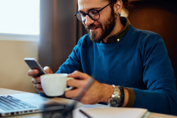 Good-looking caucasian young businessman sitting in his office, holding cup of coffee and looking at smart phone. Selective focus on smart phone.