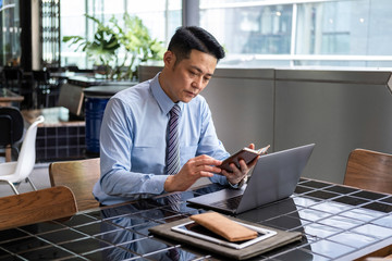 Portrait of successful businessman holding smart phone and looking at it while sitting at cafe and doing his work at laptop computer