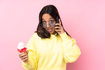 Young brunette girl holding a cornet ice cream over isolated pink background keeping a conversation...