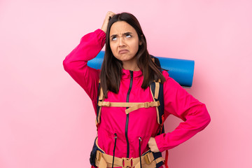 Young mountaineer girl with a big backpack over isolated pink background having doubts while scratching head