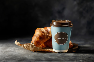 Fresh hot coffee in paper cup and tasty croissants on dark mood background. 