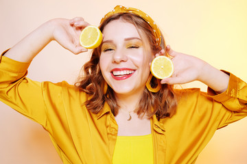 Beautiful girl, yellow make-up, bright colors. Girl holds lemons near her face, smiling. Facial masks made of lemon, procedures with lemon. Girl with accessories, big earrings, headband.