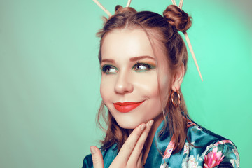 Fototapeta premium Japanese / Chinese style girl with chopsticks in her hair. Girl with an interesting hairstyle, on a green background in a Japanese, Chinese bathrobe. The girl is looking at the camera. Sushi discounts