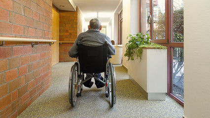 An elderly man, senior, is sitting in a wheelchair in the corridor  near the window with his back...