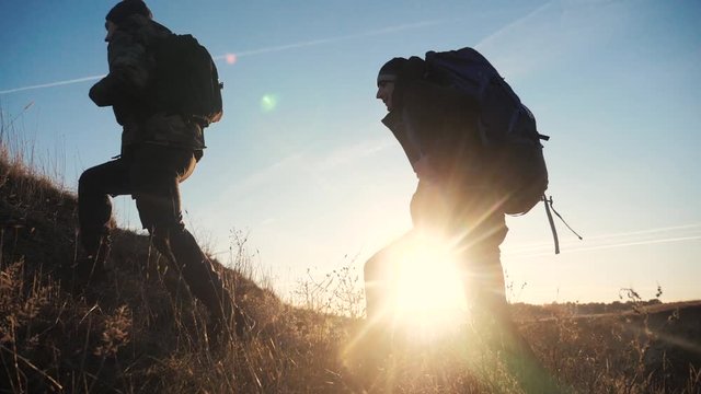 lifestyle two tourists teamwork with backpacks slow motion walking go silhouette in the sunlight glare of the sun at sunset. two men hikers climb mountains overcoming difficulties concept teamwork