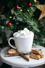 Obraz na płótnie Canvas Coffee in a white cup with marshmallows. Morning festive coffee with traditional Italian cantuccini almond cookies. A cup of coffee on a background of green fir branches on a white stand.