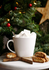 Coffee in a white cup with marshmallows. Morning festive coffee with traditional Italian cantuccini almond cookies. A cup of coffee on a background of green fir branches on a white stand.
