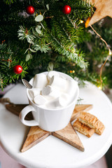 Coffee in a white cup with marshmallows. Morning festive coffee with traditional Italian cantuccini almond cookies. A cup of coffee on a background of green fir branches on a white stand.