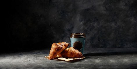 Fresh hot coffee in paper cup and tasty croissants on dark mood background. 