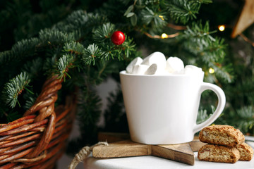 Coffee in a white cup with marshmallows. Morning festive coffee with traditional Italian cantuccini almond cookies. A cup of coffee on a background of green fir branches on a white stand.