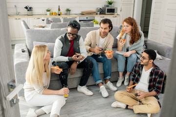 Horizontal high angle shot of young men and women spending weekend together in modern apartment eating pizza and chatting