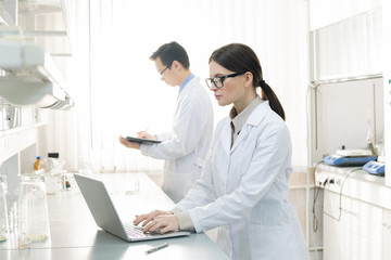 Horizontal portrait of two young laboratory scientists wearing white coats making notes in papers...