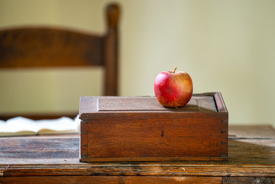 A red apple sits on a dusty teacher's desk in an antique schoolhouse.