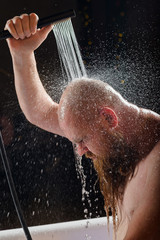 Male takes a shower. A brutal man with a red beard is standing in the bathroom under running water and washes. Spray scatter on a black background.