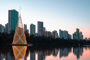 Christmas tree floating on the water of a lake and the buildings of the city on the background at the sunset. Tall tree made of christmas lights. Photo at the Igapo lake, Londrina PR Brazil.