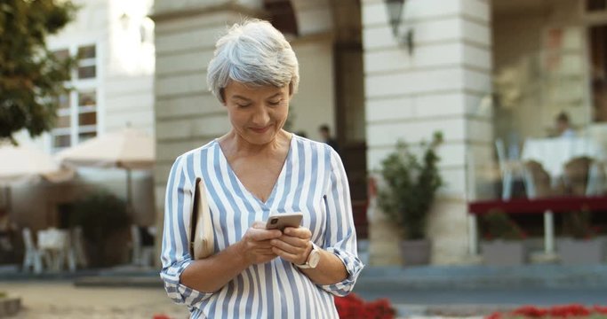 Caucasian beautiful old woman standing in center city and tapping message on smartphone. Grandmother using cellphone while typing sms. Senior nice female texting and scrolling on phone outdoors.