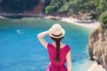 Young graceful woman at sea beach in summer vacation. Rear view of girl traveler in straw hat. Concept of travel, femininity, solo female tourism, holiday, adventure. Lady looking at ocean view.