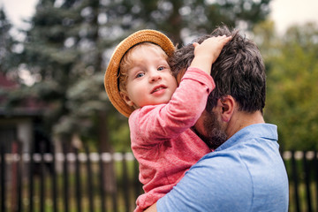 A close-up of father with toddler boy standing outdoors in garden in summer.