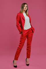 Blonde lady in red pantsuit, white blouse, high black heels. Posing standing against pink studio background. Beauty, fashion. Full length
