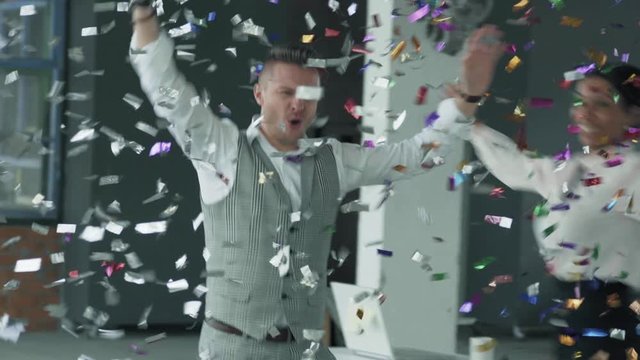 A young businessman enters the office with a reward in his hands. Colleagues join, dance with him and blow up flappers with confetti. Coworking in loft style. Office life. Winners