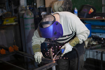 Welder at work. The welder heats the metal. Classes in the workshop. Steel processing. Welder in a blue welding mask. High melting point of metal. A man doing hard work.